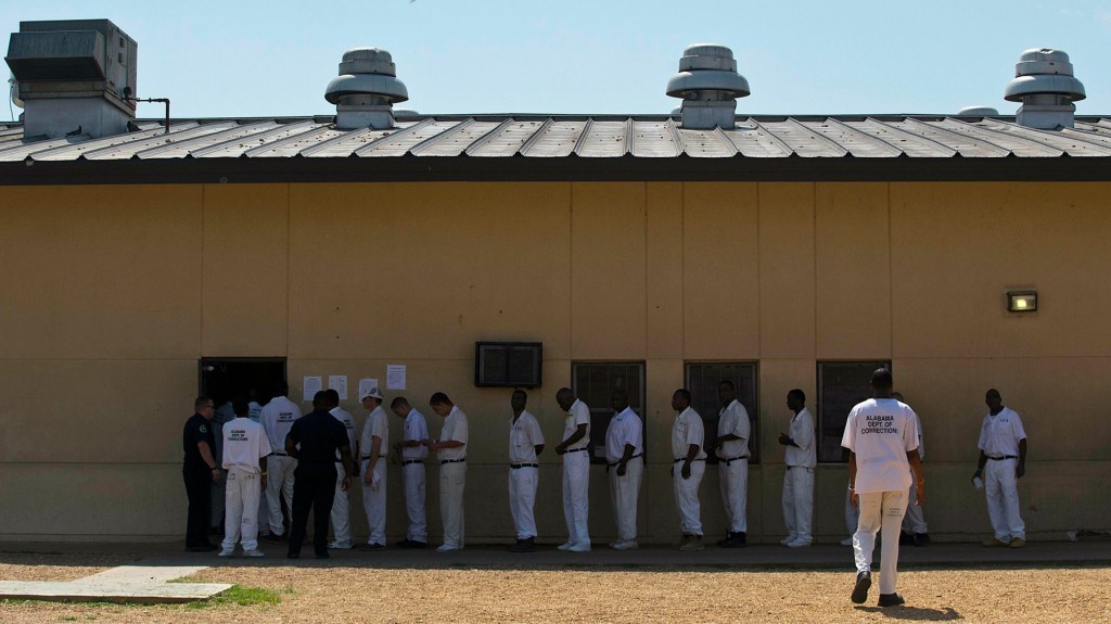 A picture of a tan building with men in white prison jumpsuits lined up in front of it. The men are predominately Black men with a couple white men in the photo. 