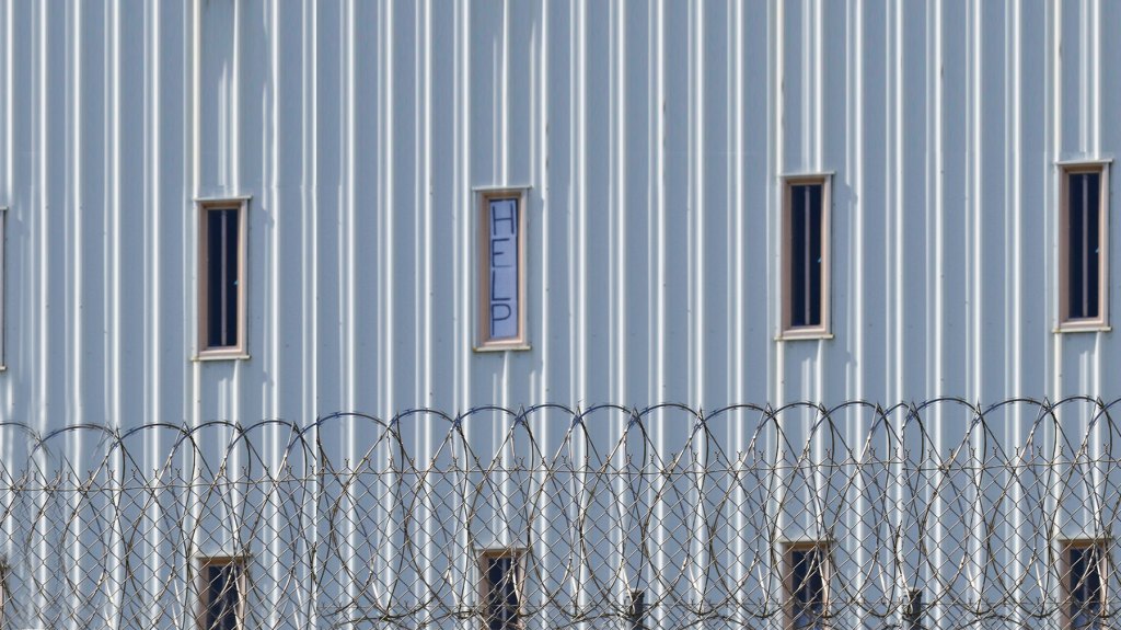 White building with chain fence in front. Windows on the building are skinny rectangles and one has a sign that reads "help". 